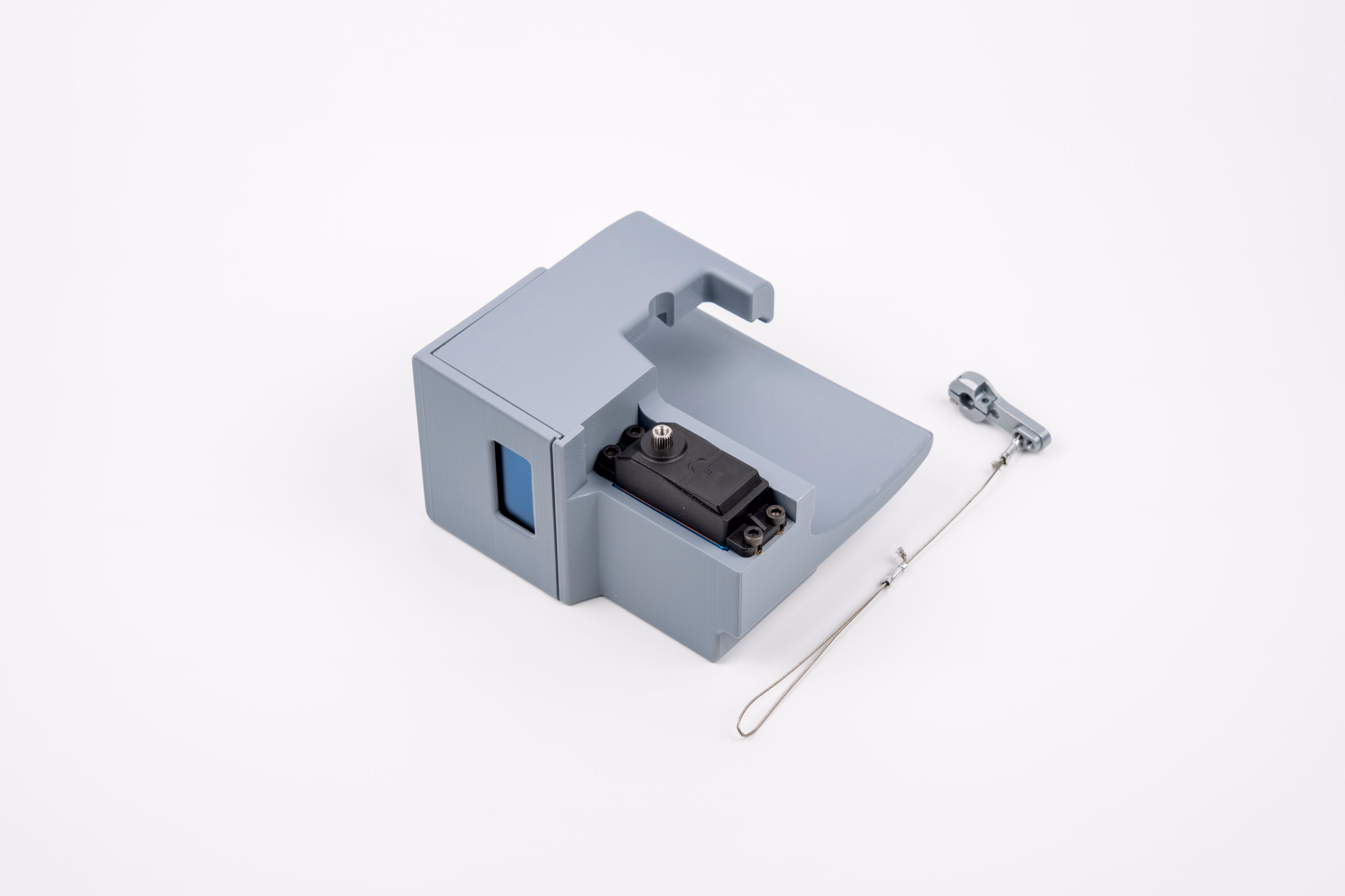 The assembled grey 3D-printed enclosure photographed on white from a three-quarter angle. A dark display window sits in the upper face; below it, a black servo motor is visible with its silver horn and a braided steel cable looping down and to the right.