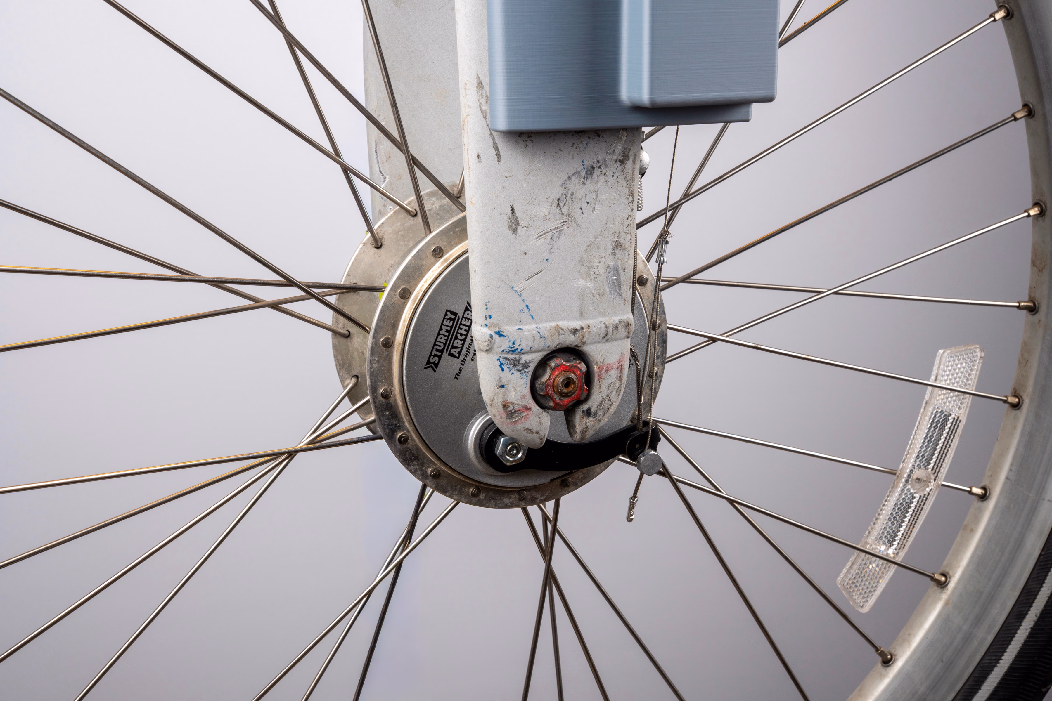 An extreme close-up of a Citi Bike's front hub, photographed through the wheel's spokes. The hub is stamped STURMEY ARCHER. A steel cable and a red brake adjuster nut are visible at the fork dropout, just below the enclosure's lower edge.