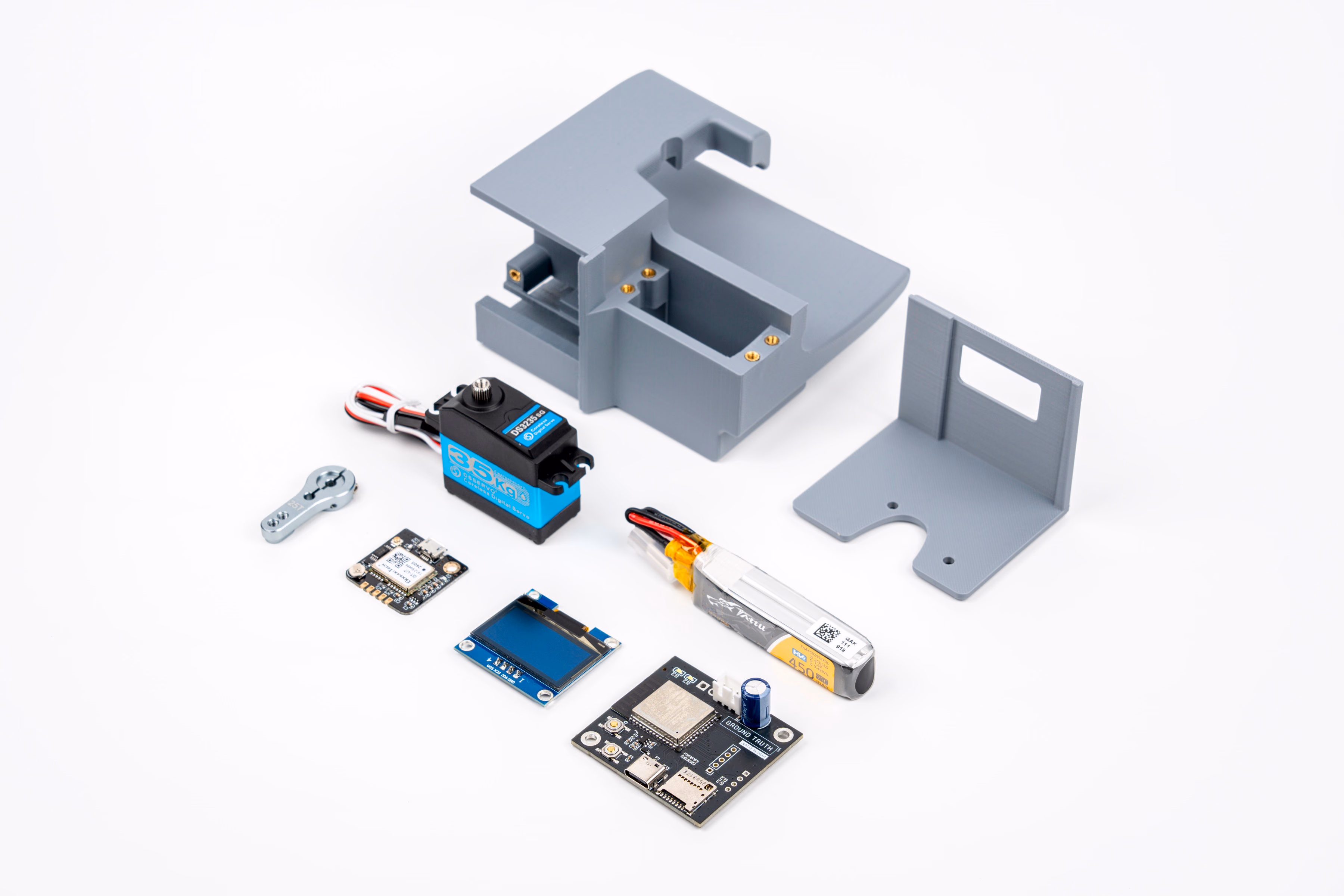 Eight components laid out on a white surface: a grey 3D-printed enclosure body with an integrated mounting hook, a separate L-shaped mounting bracket, a servo horn, a blue-and-black high-torque digital servo motor, a small GPS receiver board, a monochrome OLED display, a lithium-polymer battery pack, and a custom circuit board silkscreened GROUND TRUTH.