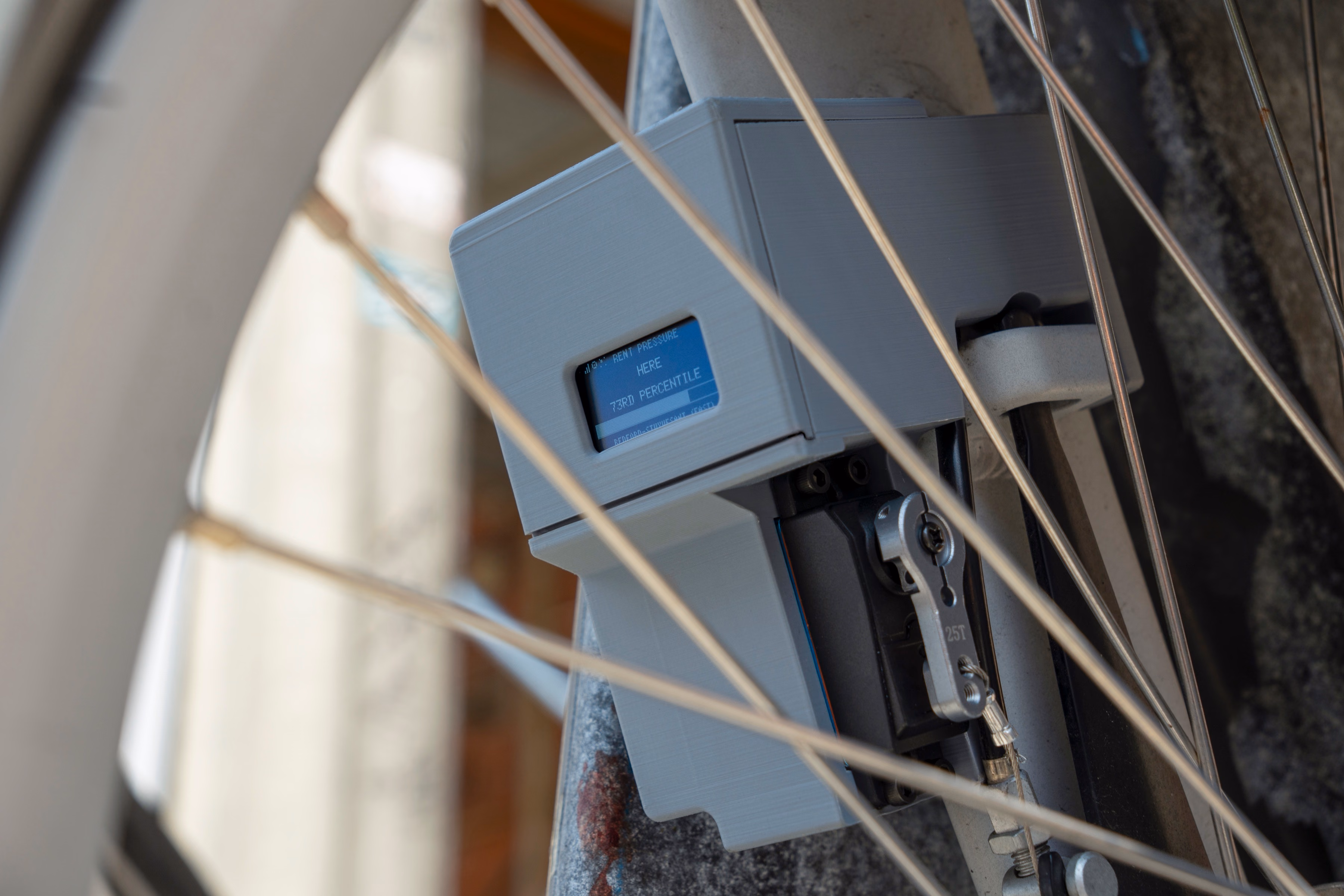 The device mounted on a Citi Bike's front fork, photographed through the wheel's spokes. Its OLED display reads RENT PRESSURE HERE, 73RD PERCENTILE, above a partially filled progress bar and a line of location text too small to read.