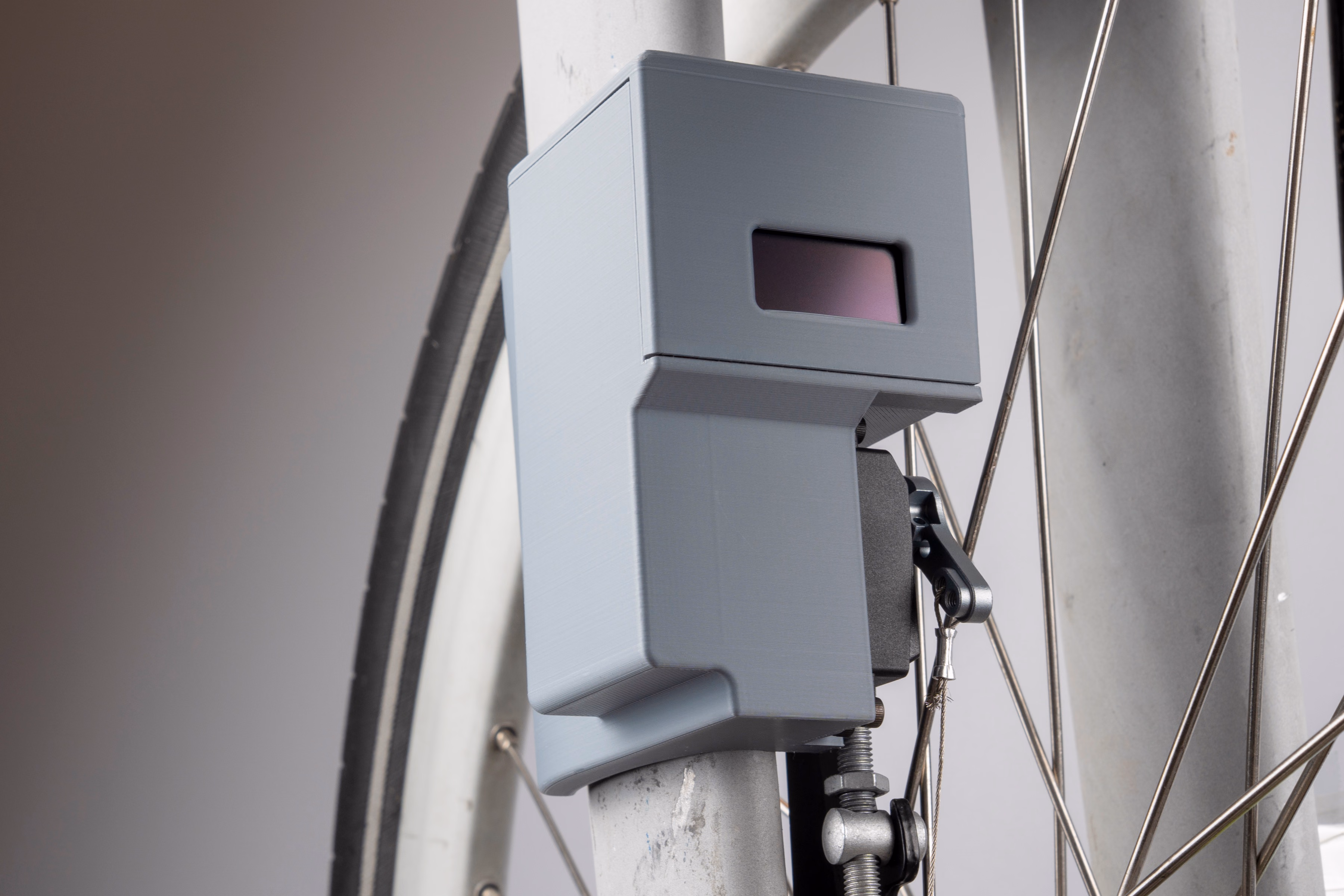 The device mounted on a Citi Bike's front fork, photographed from a three-quarter angle through the wheel's spokes. Its OLED display faces the camera, reflecting the sky; the servo and steel cable are visible below it, routed down toward the hub.