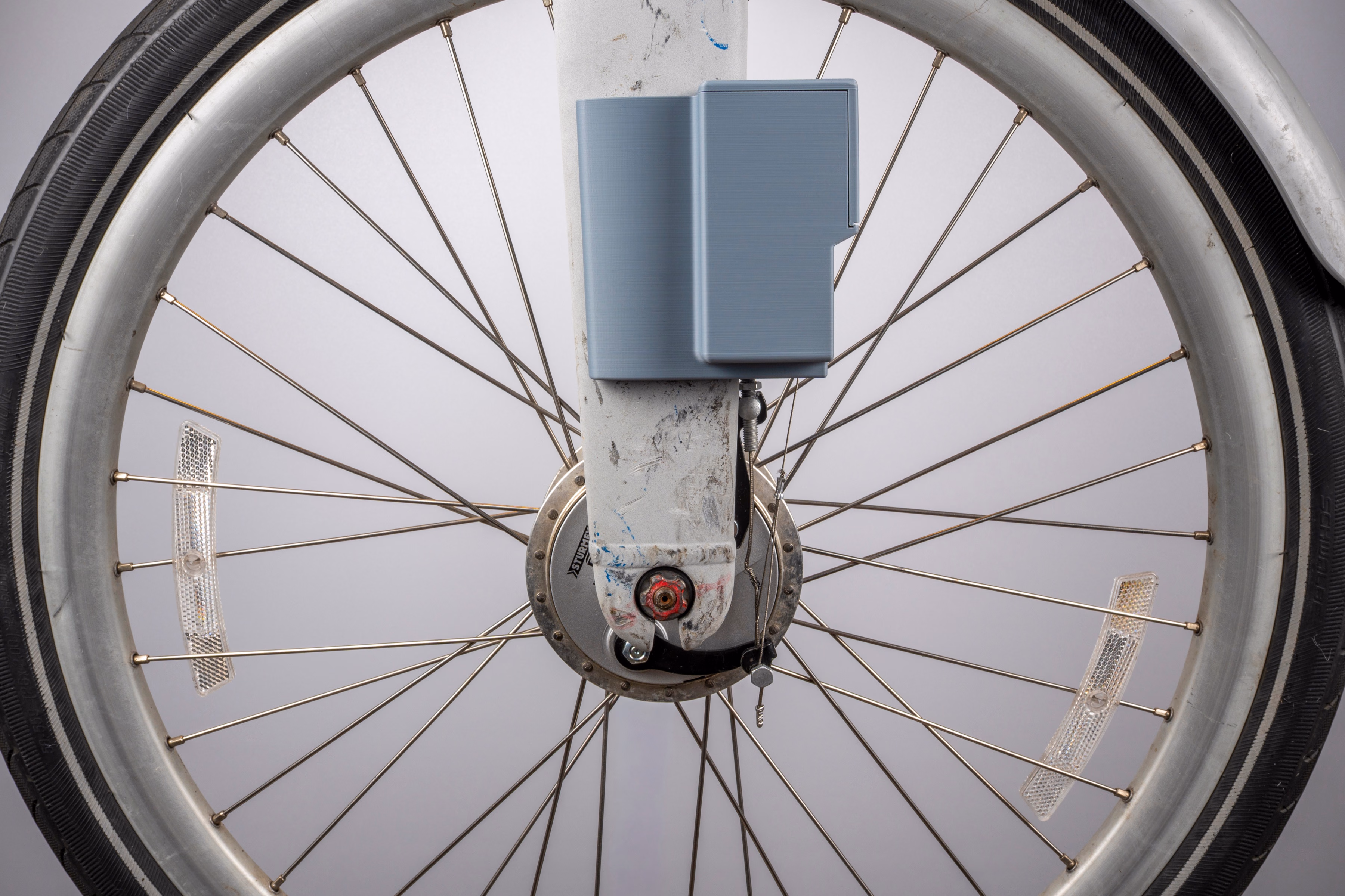 A symmetric, front-on view of a Citi Bike's front wheel. The device sits on the fork directly above the hub, centered in the frame and framed by the wheel's rim, tire, and crossing spokes.